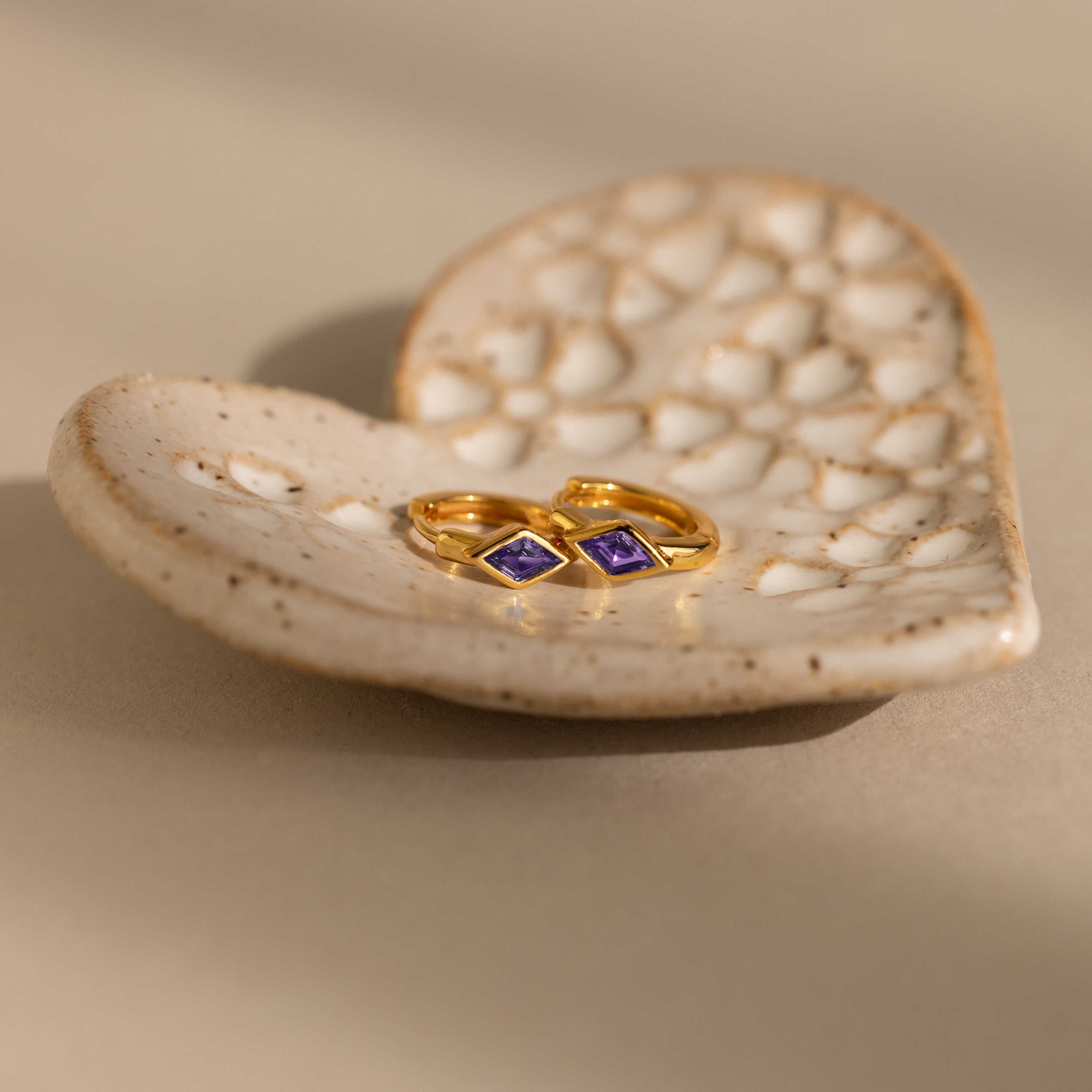 A pair of gold huggie earrings with rhombus-cut purple amethyst gemstones resting on a textured ceramic dish.