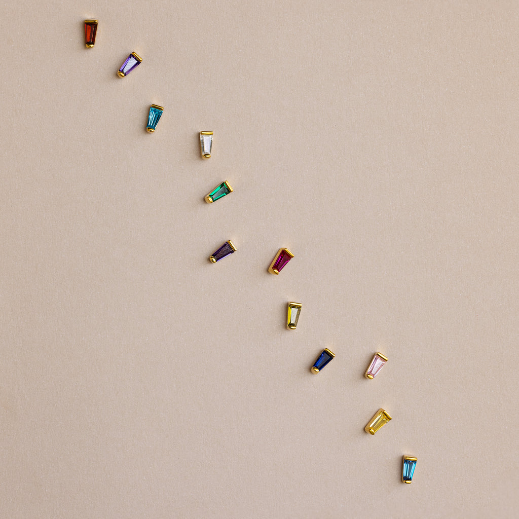 Twelve Tapered Baguette Birthstone Studs—each with a colorful gemstone—are set in gold and arranged diagonally on a beige background, highlighting the vibrant appeal of these stunning earrings.