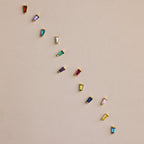 Twelve Tapered Baguette Birthstone Studs—each with a colorful gemstone—are set in gold and arranged diagonally on a beige background, highlighting the vibrant appeal of these stunning earrings.