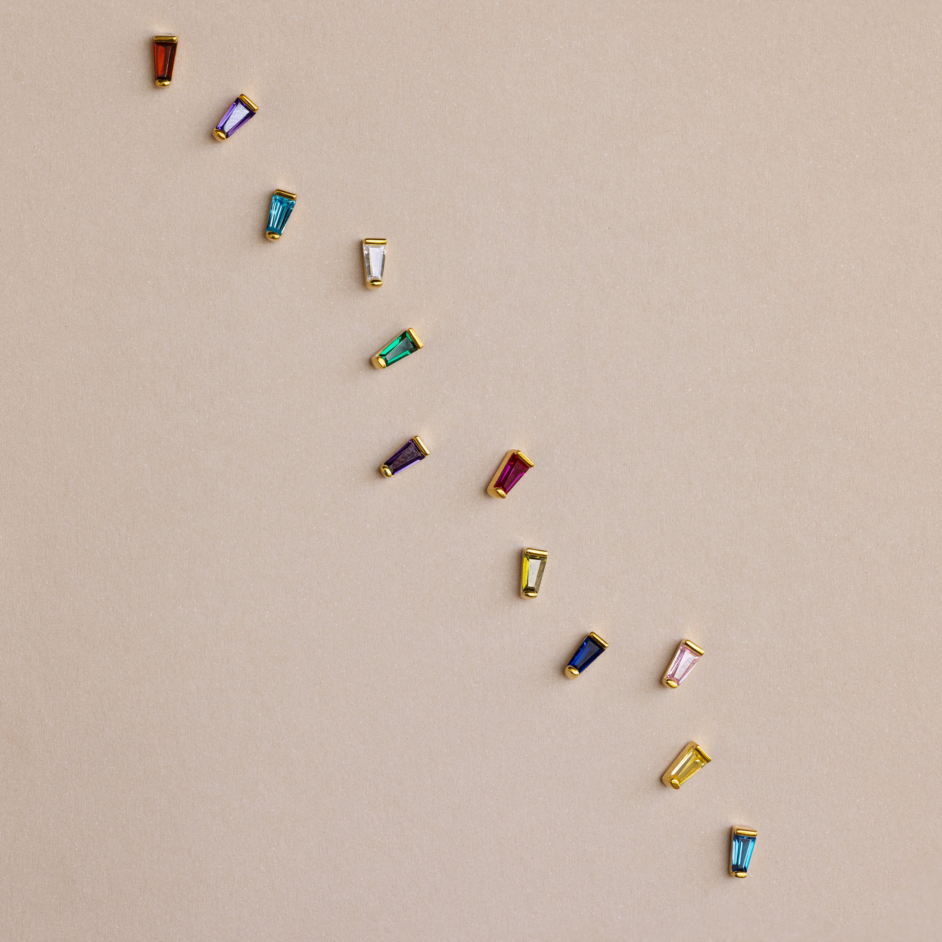 Twelve Tapered Baguette Birthstone Studs—each with a colorful gemstone—are set in gold and arranged diagonally on a beige background, highlighting the vibrant appeal of these stunning earrings.