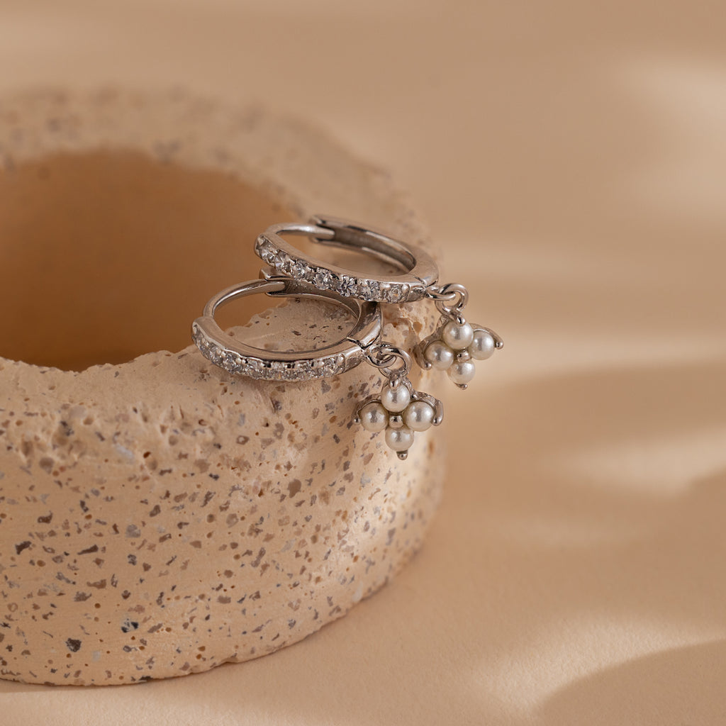 The Pearl Ivy Flower Huggies in Sterling Silver, featuring pearl cluster charms and a subtle floral design, are displayed on a textured beige ceramic surface.