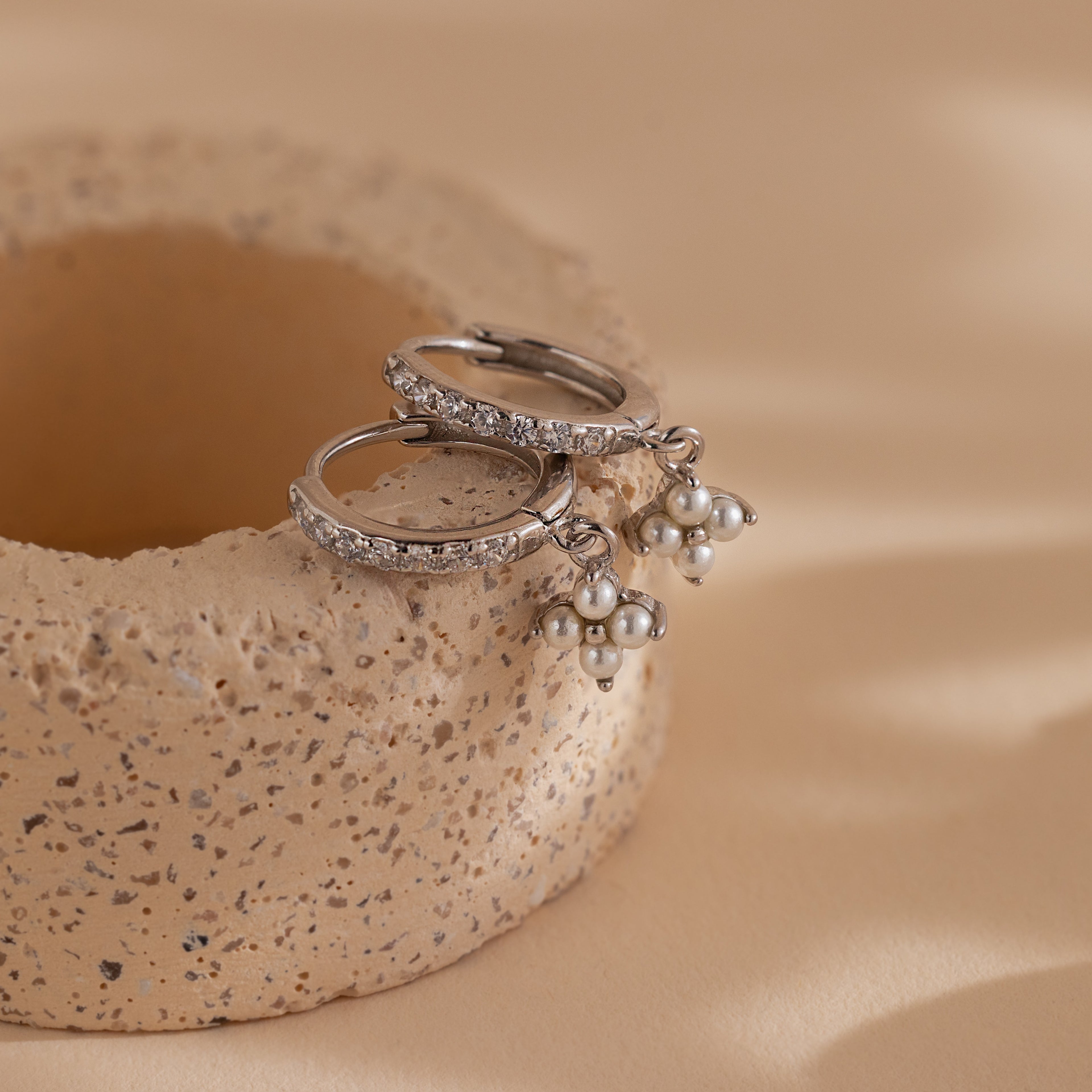 The Pearl Ivy Flower Huggies in Sterling Silver, featuring pearl cluster charms and a subtle floral design, are displayed on a textured beige ceramic surface.