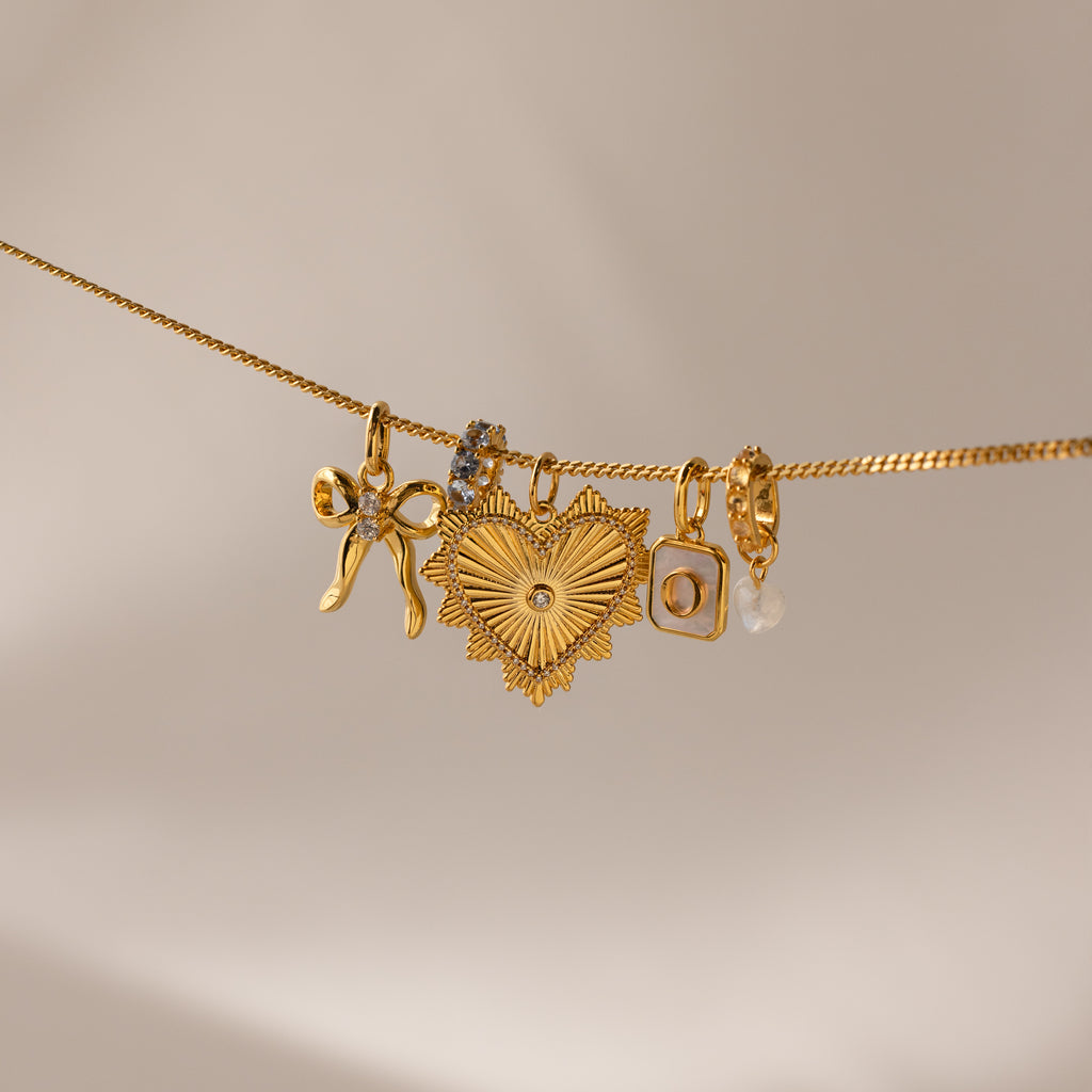 Multiple gold charms hang side by side on a gold dainty curb chain, including the gold ribbed vintage heart charm, an initial charm, heart moonstome charm, diamond infinity charm and bow charm 