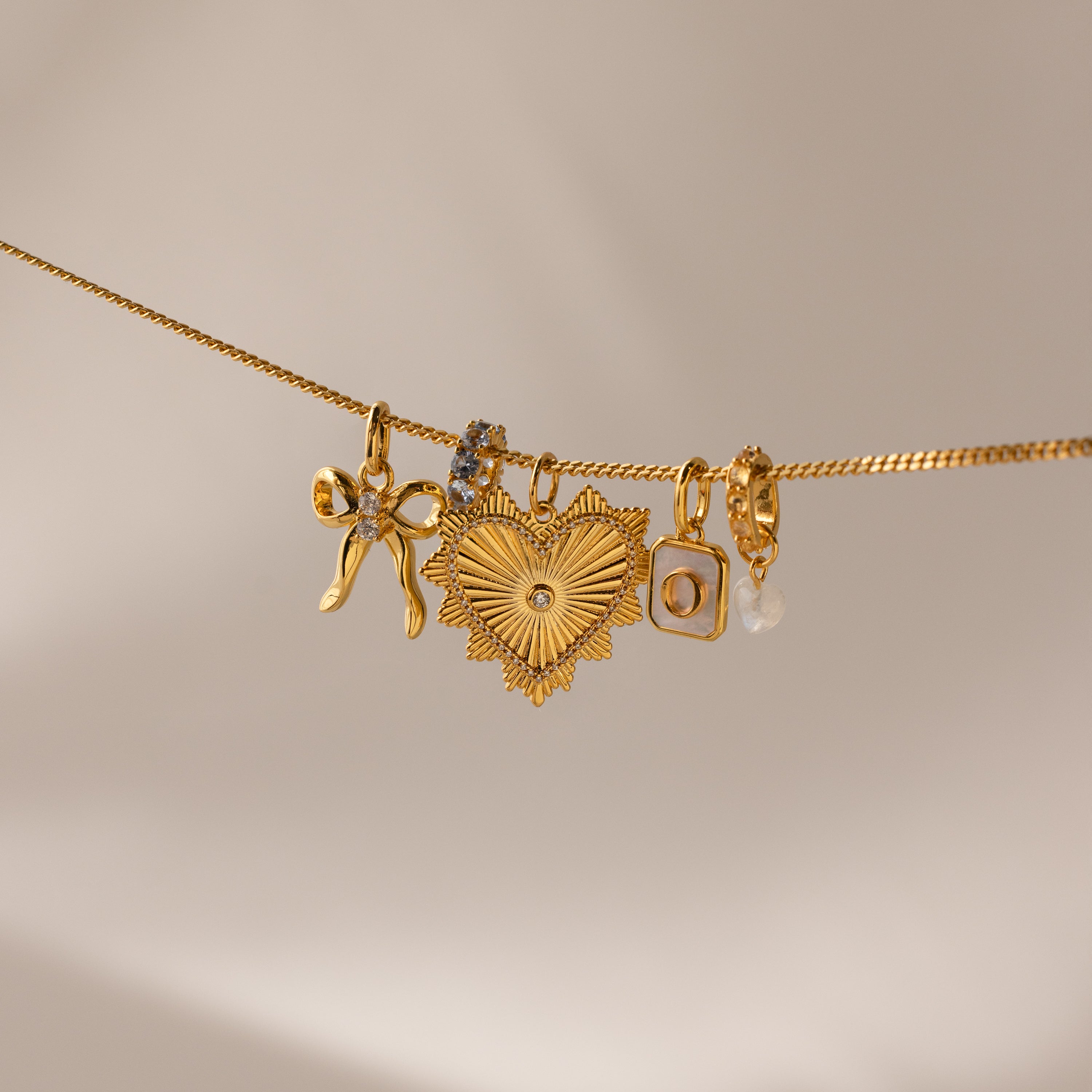 Multiple gold charms hang side by side on a gold dainty curb chain, including the gold ribbed vintage heart charm, an initial charm, heart moonstome charm, diamond infinity charm and bow charm 