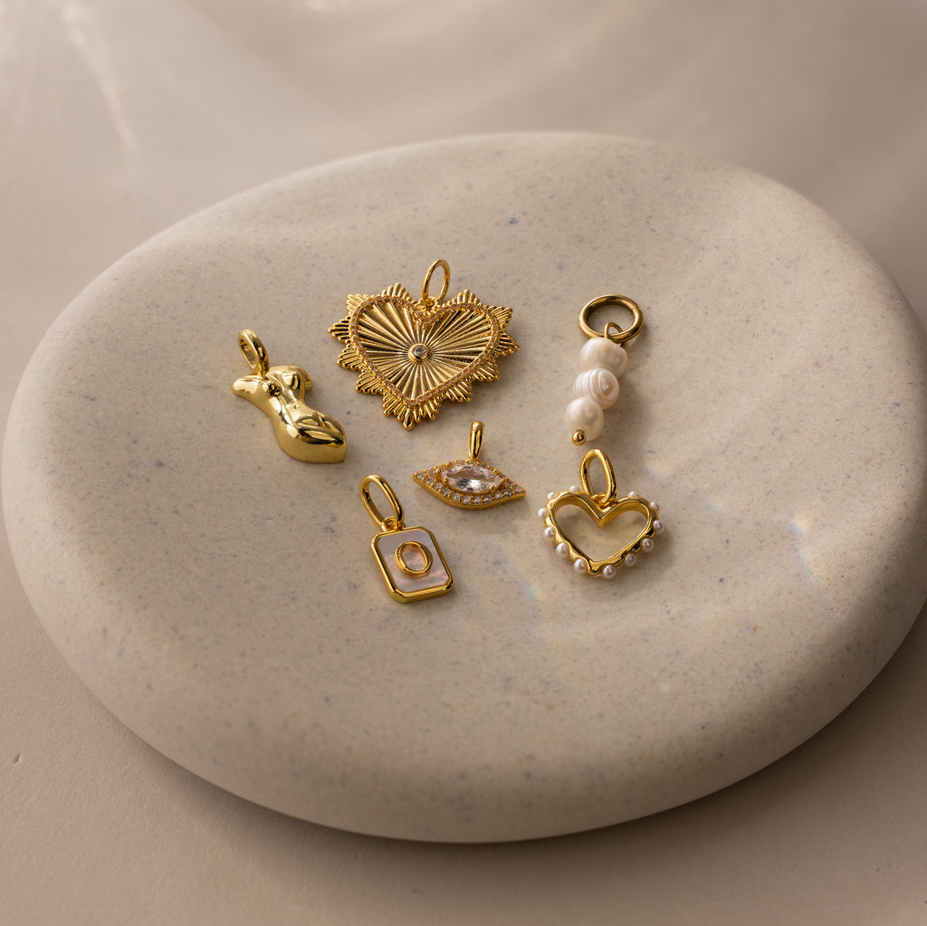 Multiple gold charms laying flat on a dish, including the vintage heart ribbed charm, a triple pearl charm, pearl heart charm, diamond marquise charm, initial charm and body charm