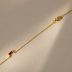 The Initial Baguette Birthstone Necklace, featuring a delicate gold chain and a small baguette-cut birthstone, is shown on a beige background in soft light.