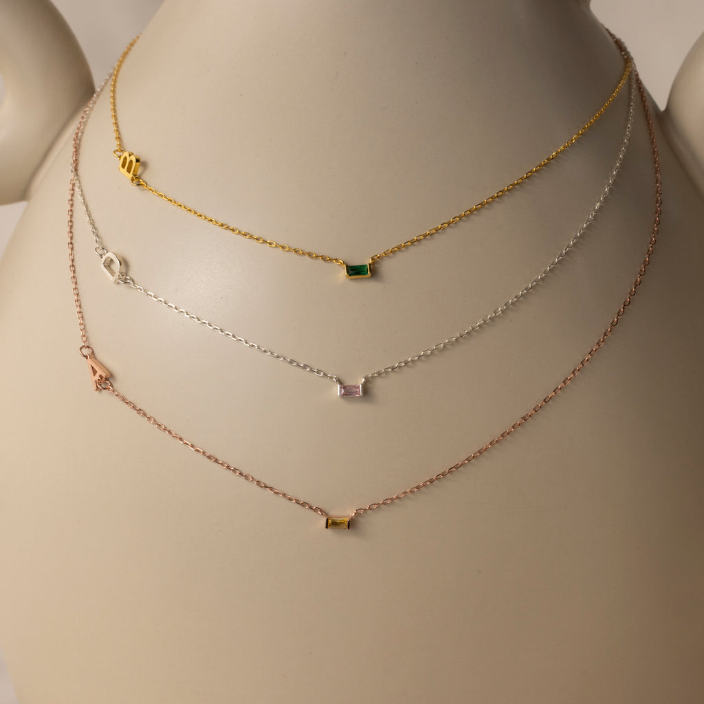 The Initial Baguette Birthstone Necklace—available in gold, silver, and rose gold—is showcased on a white mannequin bust.