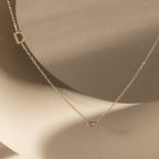 The Initial Baguette Birthstone Necklace features a gold chain with a sideways letter D charm and a small rectangular pink baguette-cut birthstone, shown on a smooth beige background.