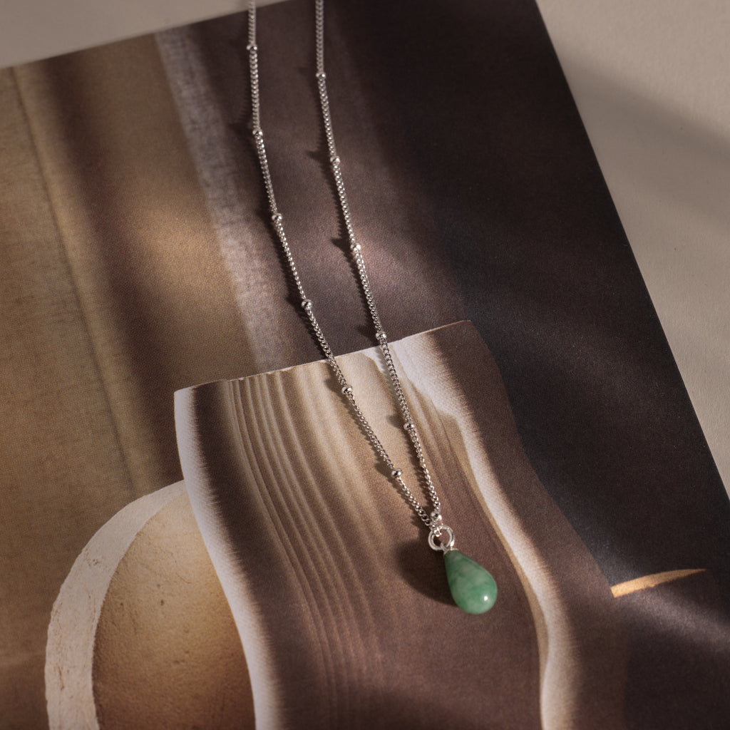 Flat-lay of a silver satellite chain necklace featuring a single green jade teardrop gemstone pendant.