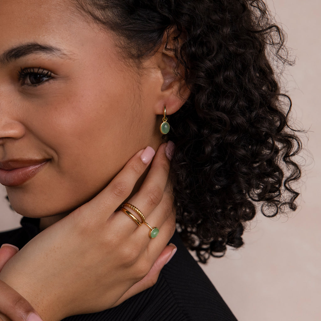 Woman with curly hair, smiling softly and touching her face and neck, wears gold jewelry featuring the Jade Drop Hoops adorned with green stones.