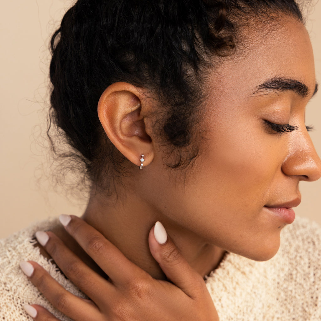 Profile view of a woman with eyes closed and a relaxed expression, wearing a silver opal and diamond huggie earring and a cozy neutral-toned sweater