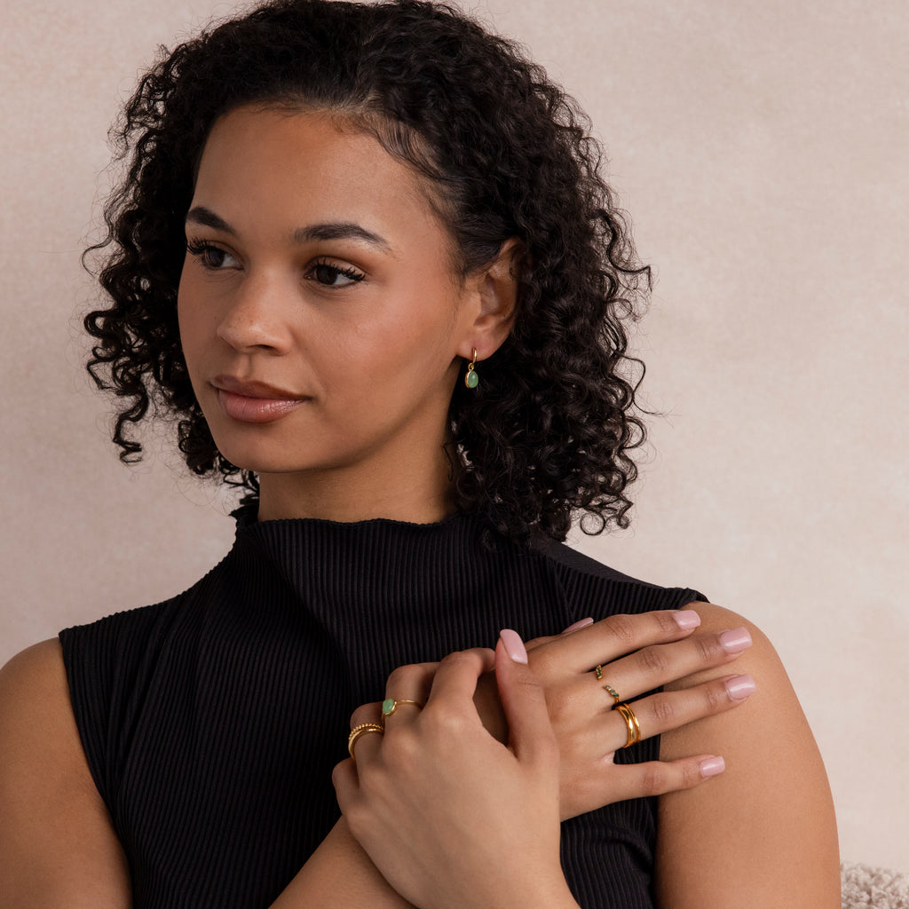 A woman in a black top models the Jade Drop Hoops, hands crossed over her chest and gazing to the side, with gold rings accenting her look.