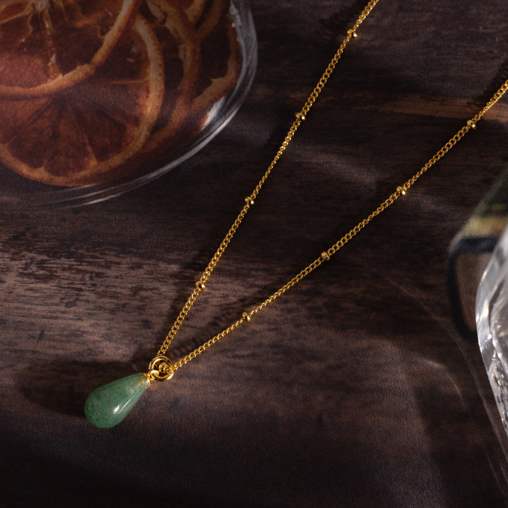 The Jade Drop Necklace in 18K Gold features a green teardrop jade pendant on a gold beaded chain, shown on a wooden surface next to dried orange slices.