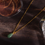 The Jade Drop Necklace in 18K Gold features a green teardrop jade pendant on a gold beaded chain, shown on a wooden surface next to dried orange slices.