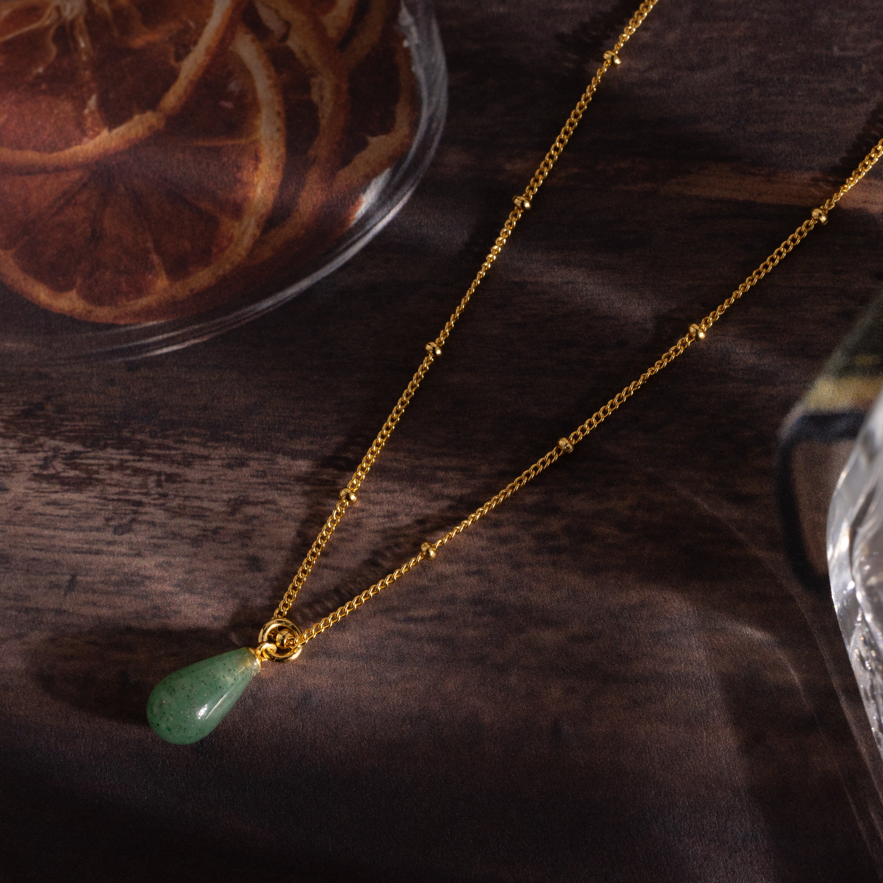 The Jade Drop Necklace in 18K Gold features a green teardrop jade pendant on a gold beaded chain, shown on a wooden surface next to dried orange slices.