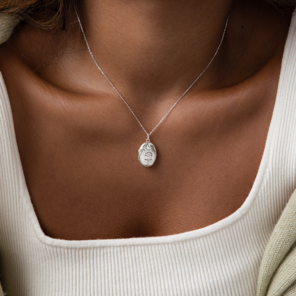 A woman wears the Floral Handwriting Locket Necklace in Sterling Silver over a white ribbed top, shown from shoulders to chest.