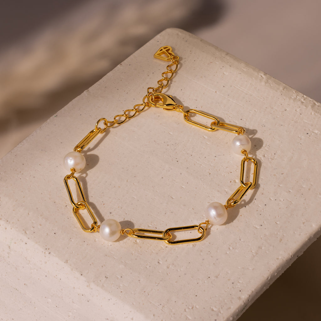The Pearl Station Paperclip Bracelet features pearl accents on a gold paperclip chain, displayed on a textured beige surface.