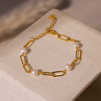 The Pearl Station Paperclip Bracelet features pearl accents on a gold paperclip chain, displayed on a textured beige surface.