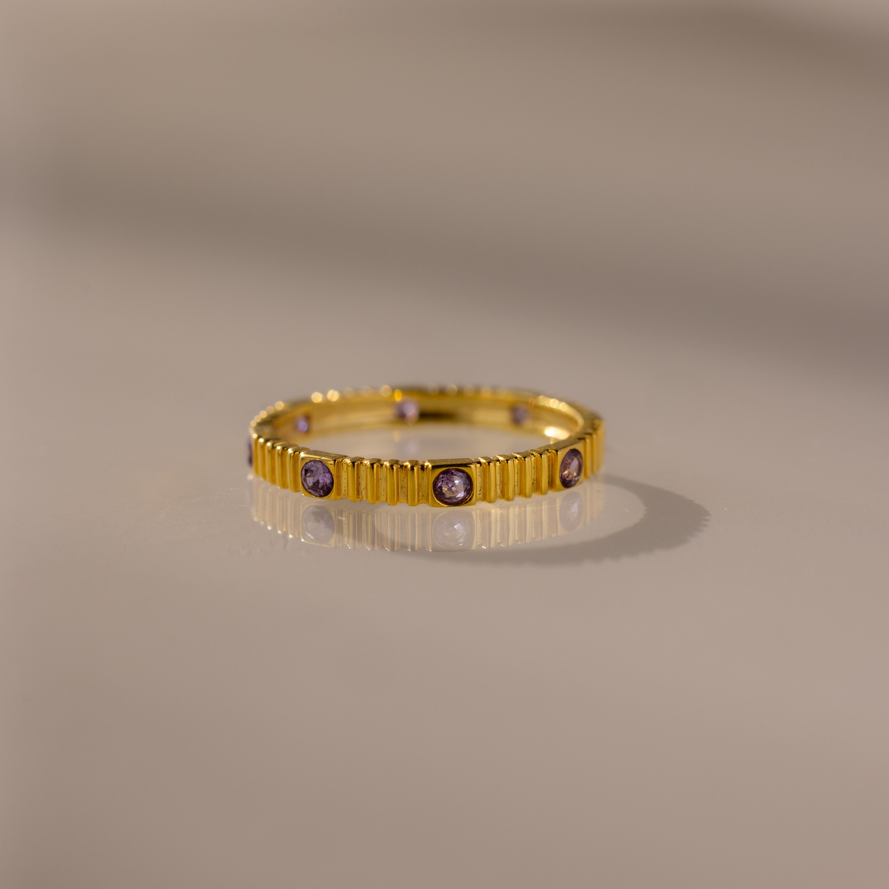 The Ribbed Round Birthstone Eternity Ring in Alexandrite (June) features vertical ridges and small purple gemstones, displayed on a smooth, neutral surface.