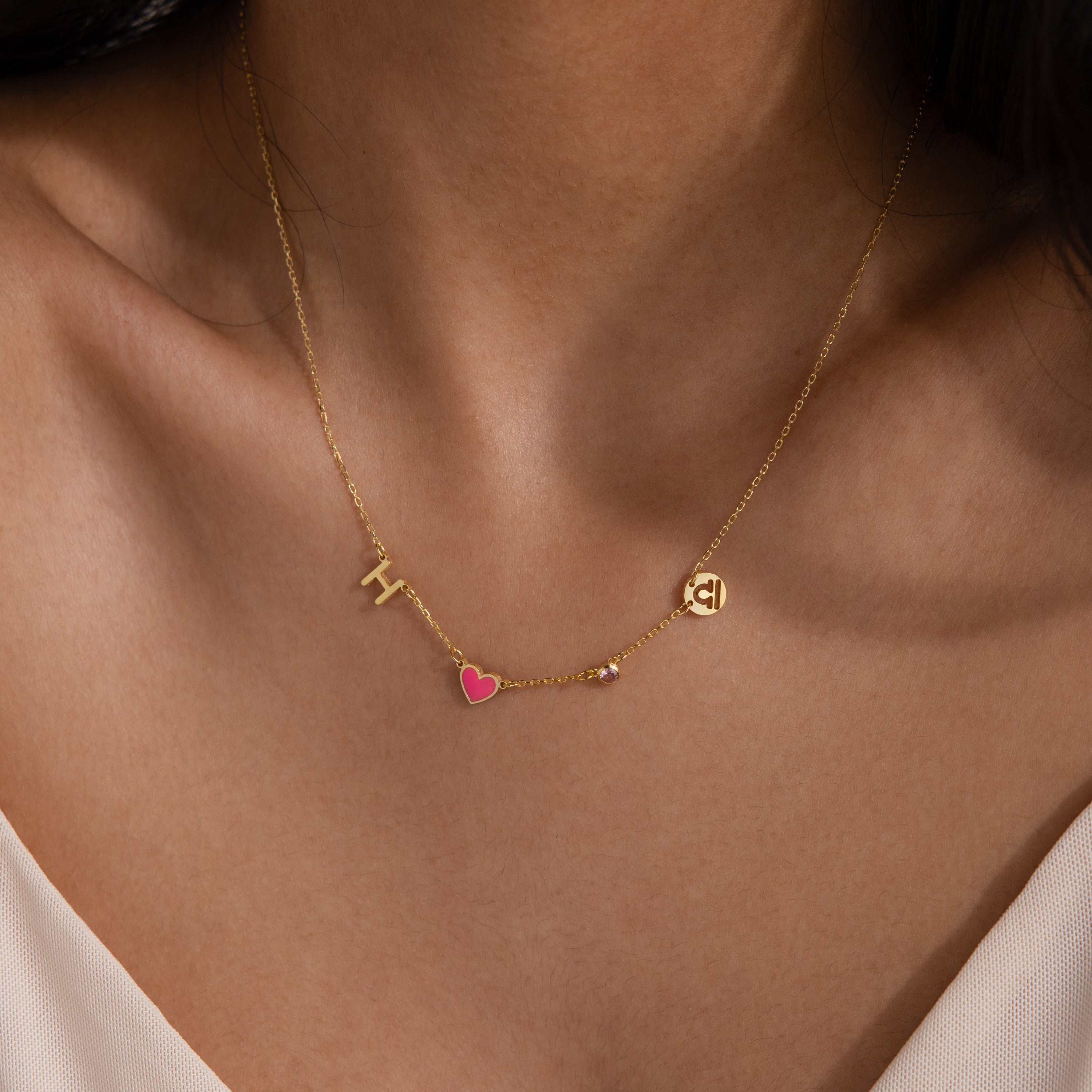 The Dainty Space Charm Necklace features gold letter charms H and C, plus a pink heart charm, worn with a white top—an ideal customizable necklace for any occasion.