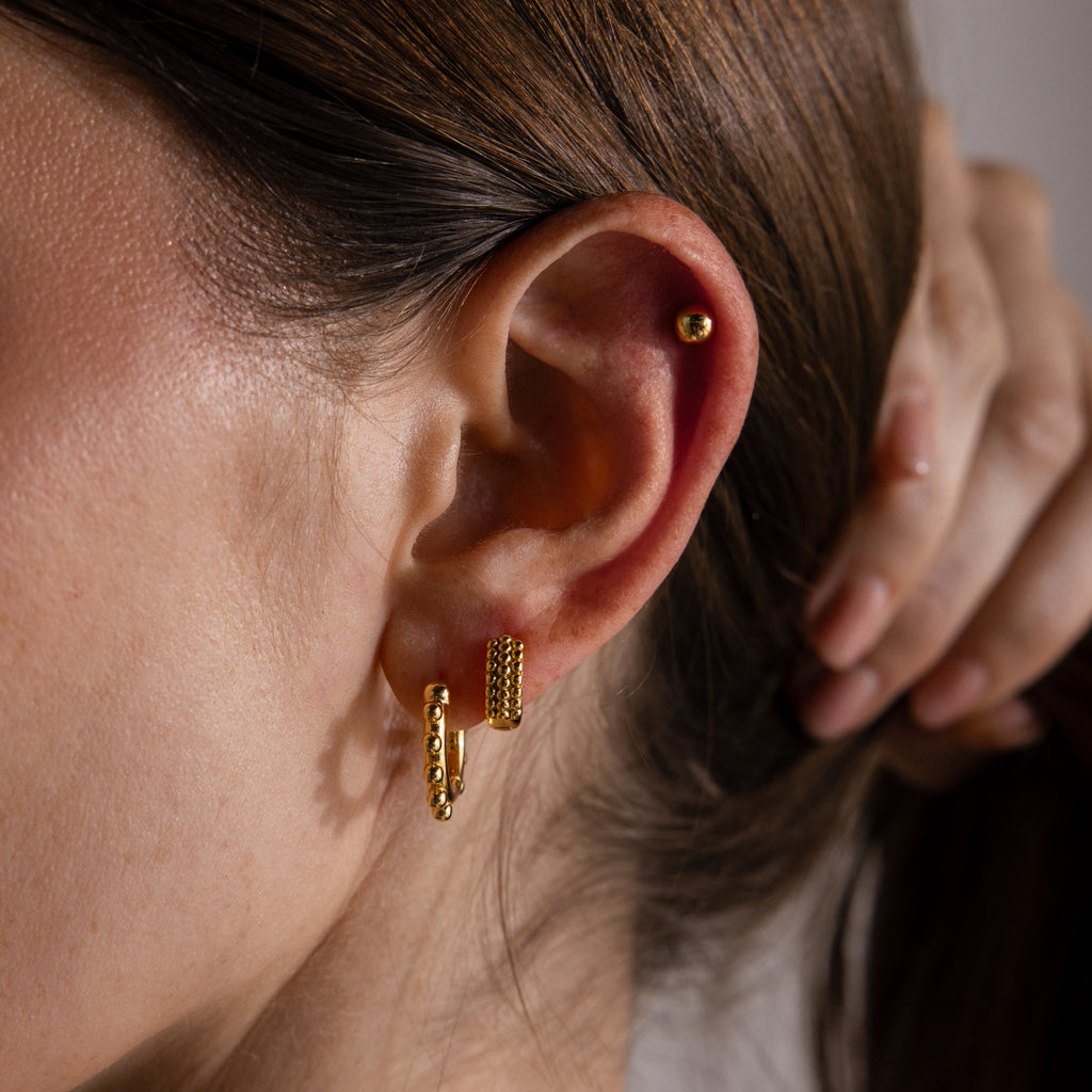 Close-up of a woman's ear wearing three gold hoop earrings, featuring the Beaded Hoops in 18K Gold, as her hand softly touches her hair in the background.