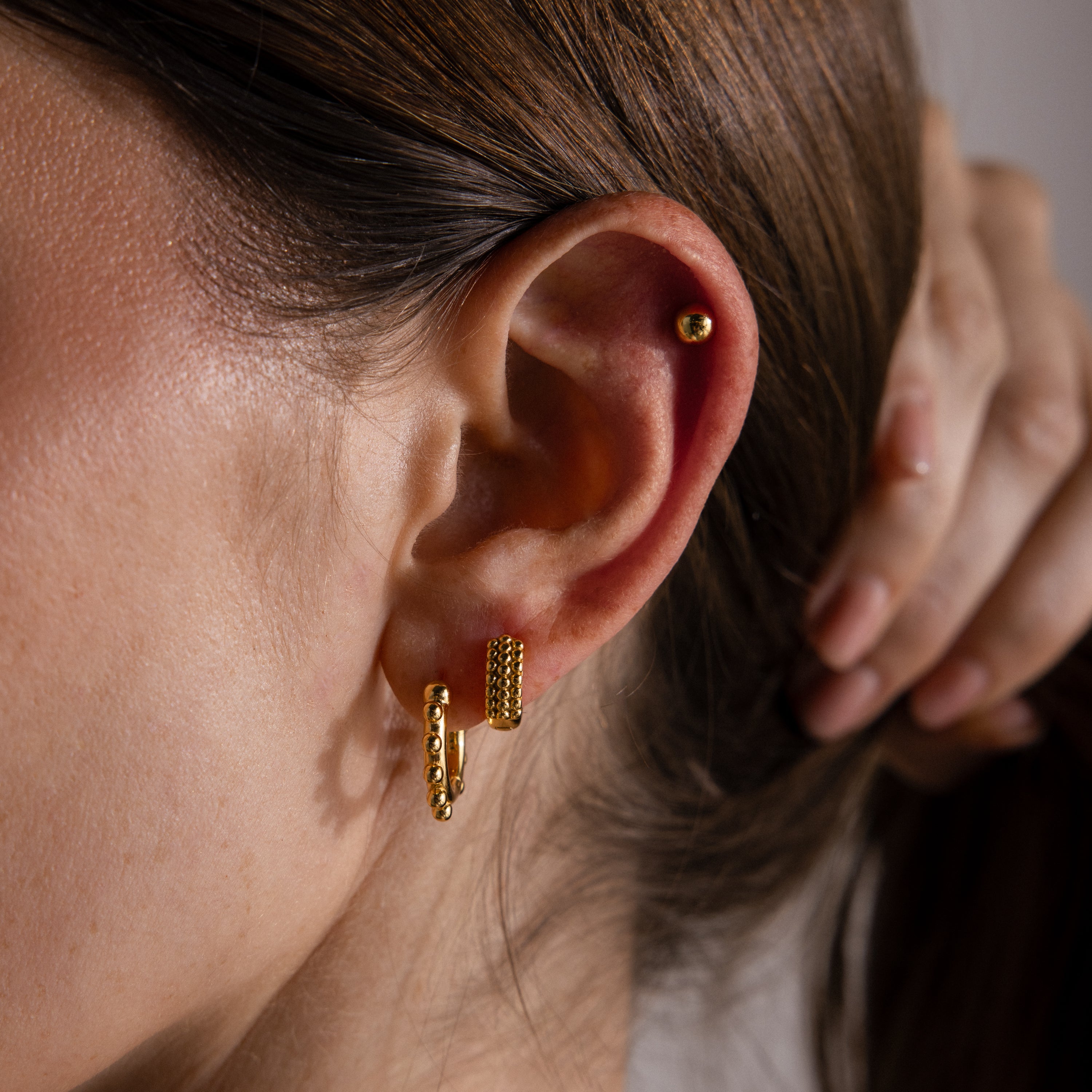 A close-up showcases a woman's ear wearing Beaded Hoops and two other gold earrings, as her hand gently touches her brown hair in the background.