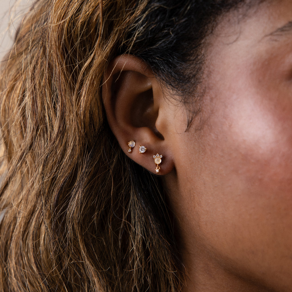 A close-up of a woman's ear showcases four Halo Moonstone Drop Studs in 18K Gold, exuding royal elegance along the earlobe.