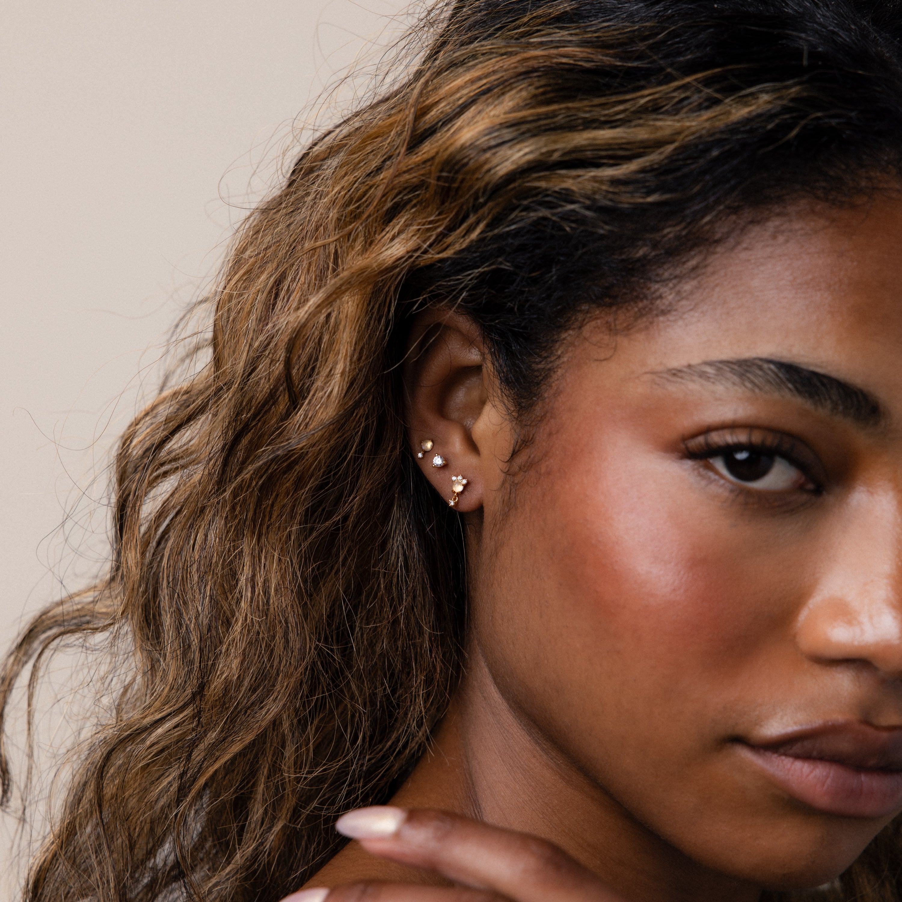 Woman with wavy hair wears Halo Moonstone Drop Studs in 18K Gold, gazing at the camera against a neutral background and exuding royal elegance.