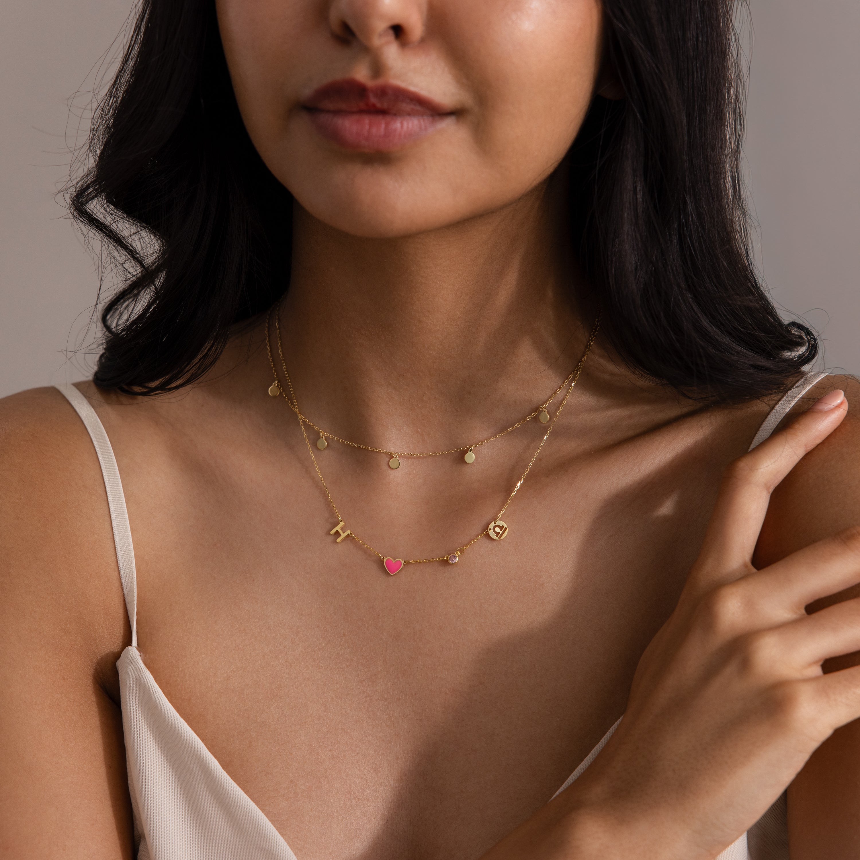 A woman in a cream spaghetti strap top showcases the Dainty Space Charm Necklace, featuring customizable small gold pendants and a pink heart.