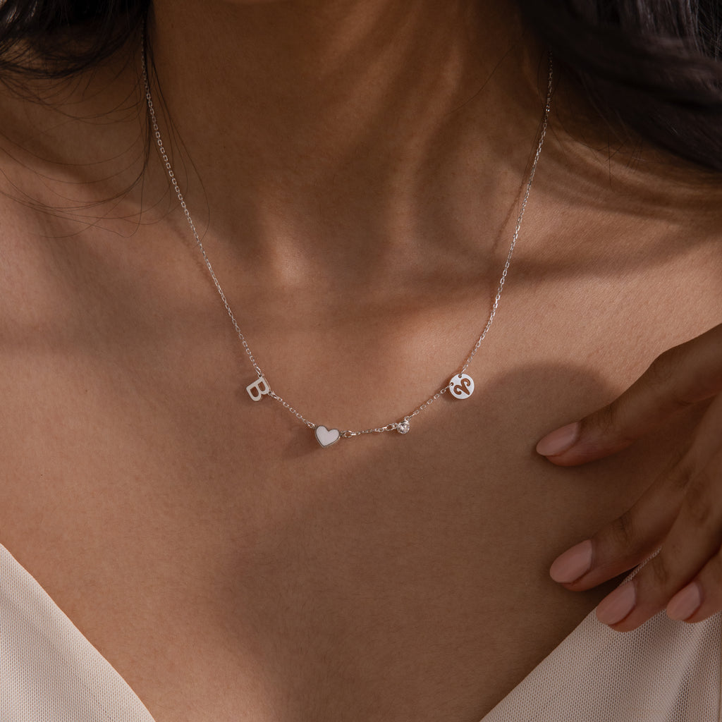 A woman wears the Dainty Space Charm Necklace in Sterling Silver, featuring personalized letter charms, a heart, and an ampersand on her bare neckline.
