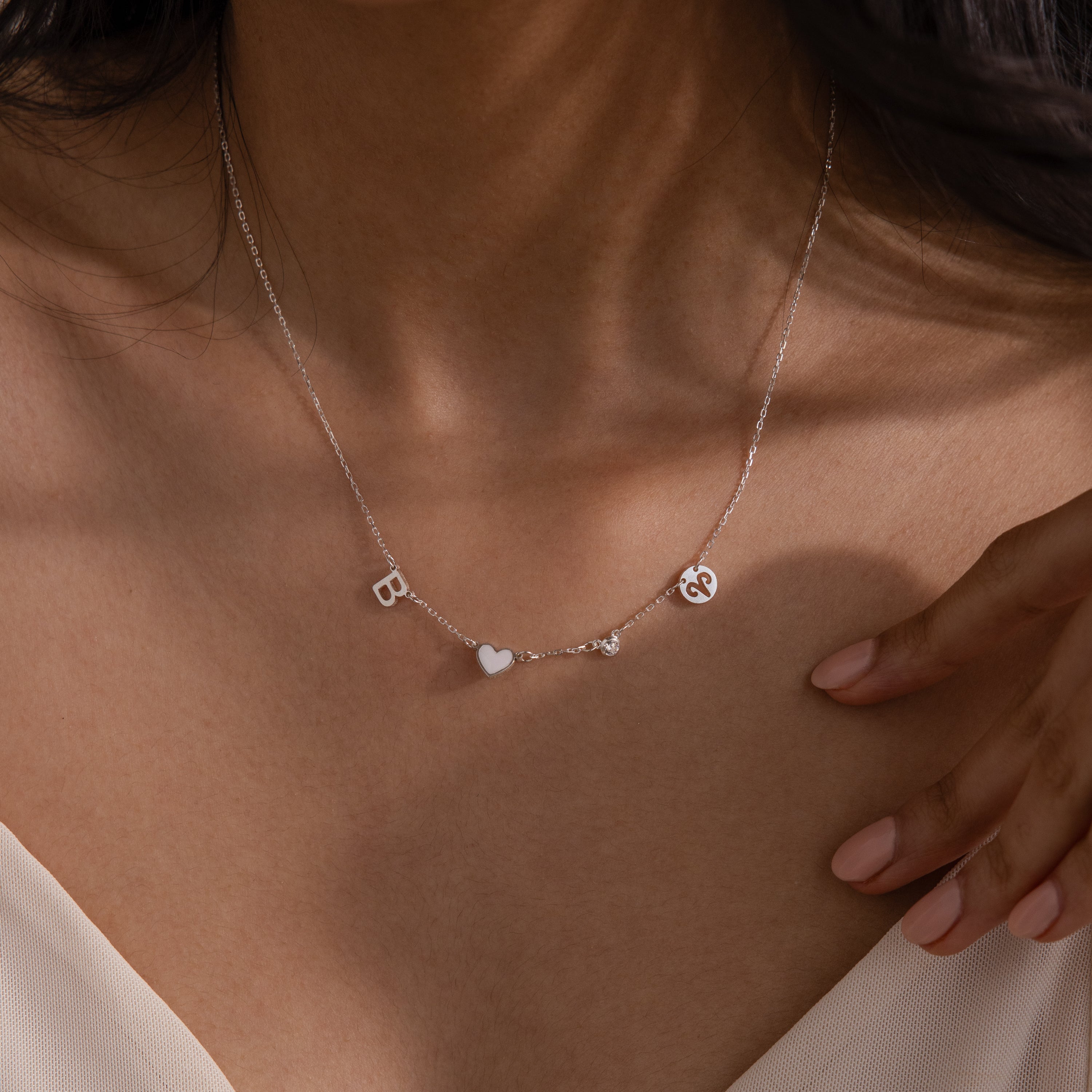 A woman wears the Dainty Space Charm Necklace in Sterling Silver, featuring personalized letter charms, a heart, and an ampersand on her bare neckline.