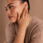A woman with sleek dark hair wears a fuzzy beige sweater and the Love Knot Bracelet in 18K Gold, touching her ear—an elegant double chain piece that's a perfect gift for couples.