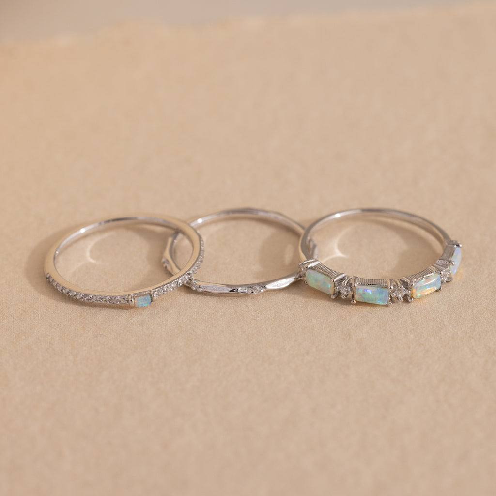 The Pave Opal Ring Set in Sterling Silver features three delicate bands with opal and diamond accents, arranged on a light beige surface—a timeless trio perfect for stacking or gifting.
