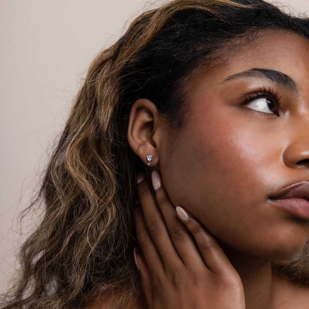 A woman with wavy hair touches her neck, displaying Halo Moonstone Drop Studs in Sterling Silver and natural makeup, radiating royal elegance.