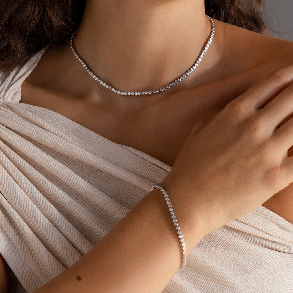 A woman wears a white off-shoulder top, displaying the Diamond Bezel Tennis Necklace & Bracelet Set in White Gold—an elegant, unforgettable anniversary gift.