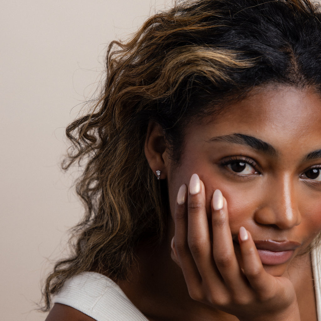 A woman with wavy hair gazes thoughtfully at the camera, resting her face on her hand as Halo Moonstone Drop Studs in Sterling Silver add a touch of royal elegance to her serene pose.