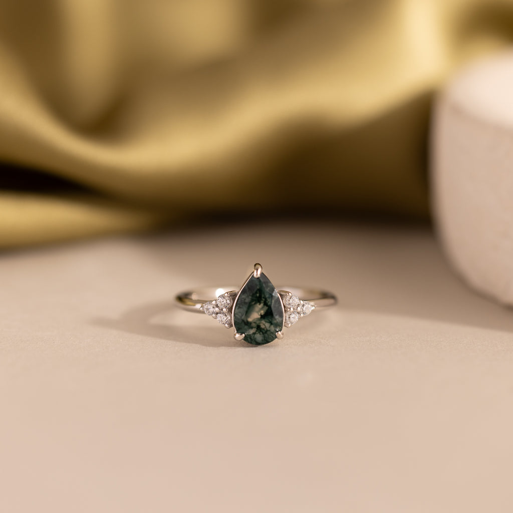 The Agate Teardrop Ring in Sterling Silver showcases a dark green gemstone with clear side stones, set in silver and displayed on a beige surface—an ideal moss agate cocktail ring for any occasion.