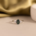 The Agate Teardrop Ring in Sterling Silver showcases a dark green gemstone with clear side stones, set in silver and displayed on a beige surface—an ideal moss agate cocktail ring for any occasion.