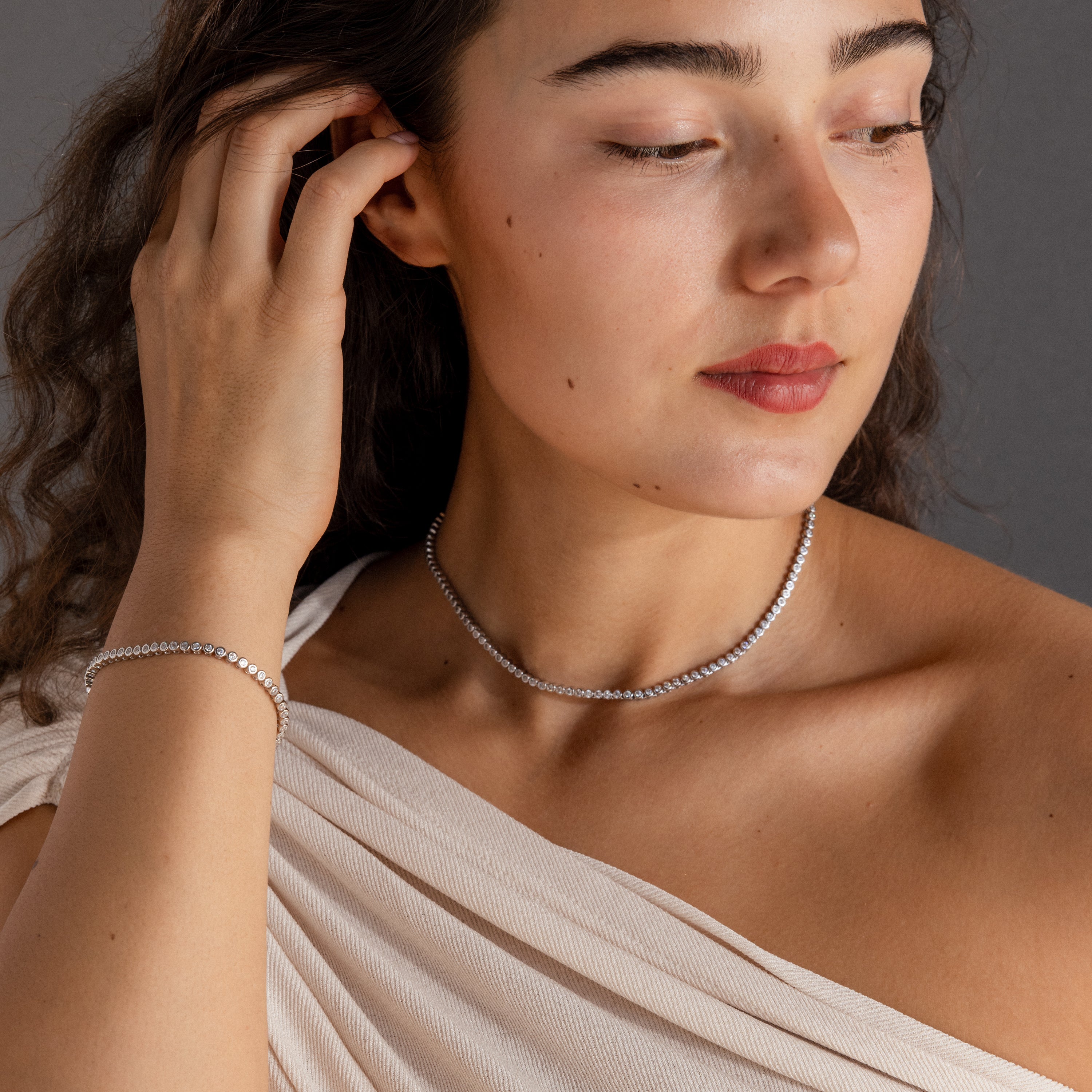 A woman wearing the Diamond Bezel Tennis Necklace & Bracelet Set touches her hair while looking down, pairing it with an off-shoulder top.