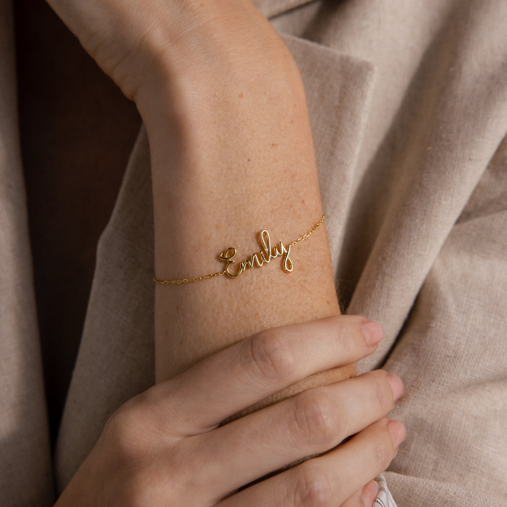 A Cursive Name Bracelet in Rose Gold spelling "Emily" decorates a wrist, the hand gently resting on an arm—a stunning personalized accessory.