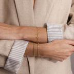 A person in a beige blazer with folded sleeves wears two gold bracelets, including the Cursive Name Bracelet in Rose Gold featuring a scripted pendant.
