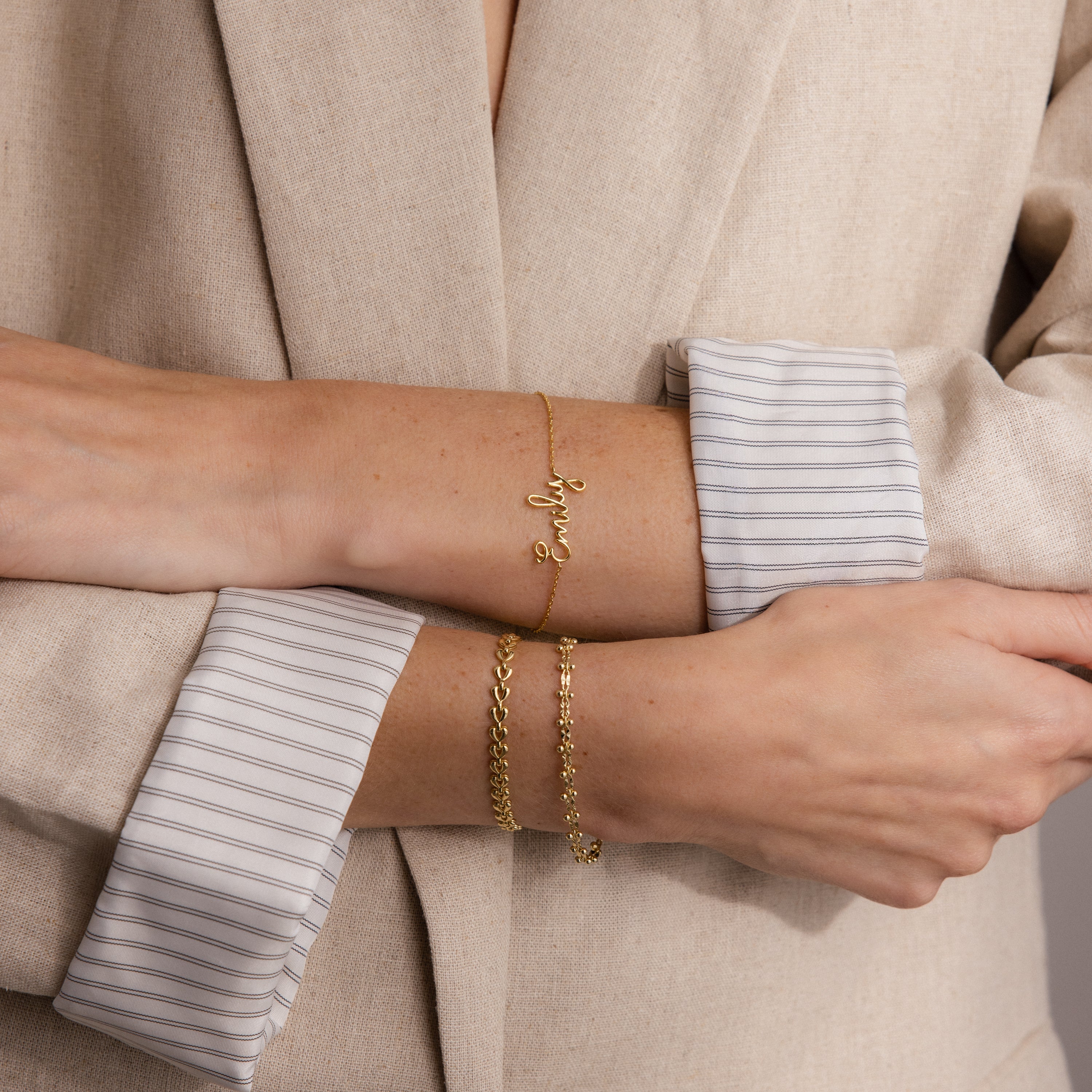 A person in a beige blazer with folded sleeves wears two gold bracelets, including the Cursive Name Bracelet in Rose Gold featuring a scripted pendant.