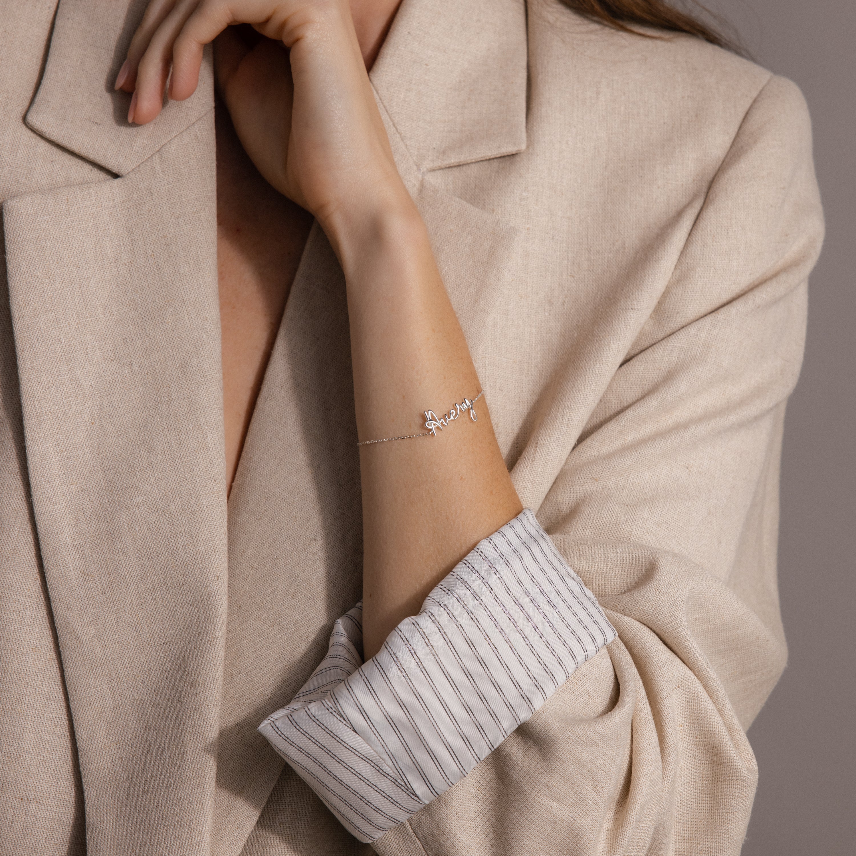 A woman in a beige blazer with striped cuffs wears the Cursive Name Bracelet, adding a personalized touch to her look.