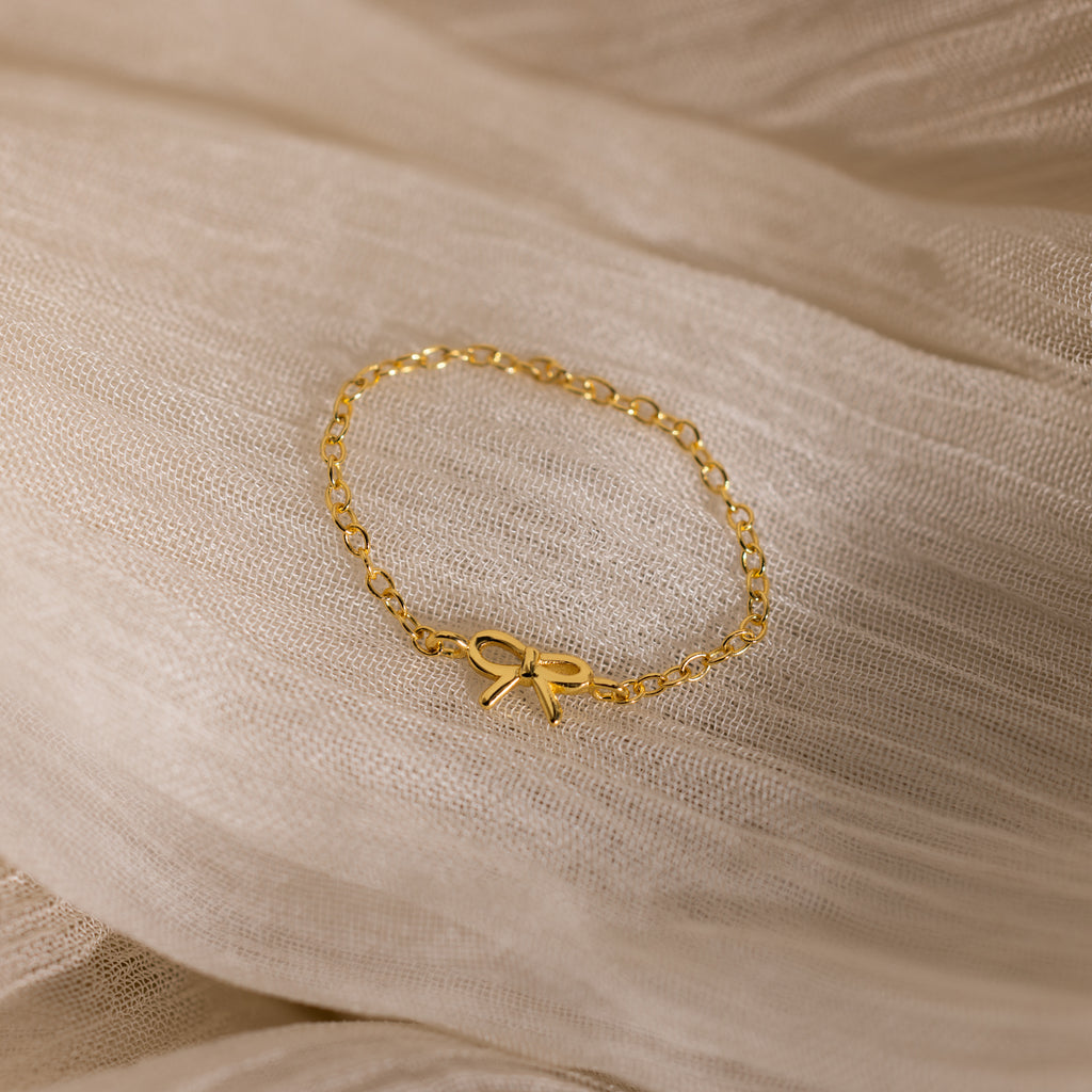 A delicate gold chain ring with a small bow centerpiece displayed on a soft, sheer beige fabric.