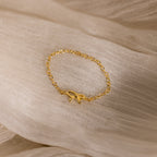 A delicate gold chain ring with a small bow centerpiece displayed on a soft, sheer beige fabric.