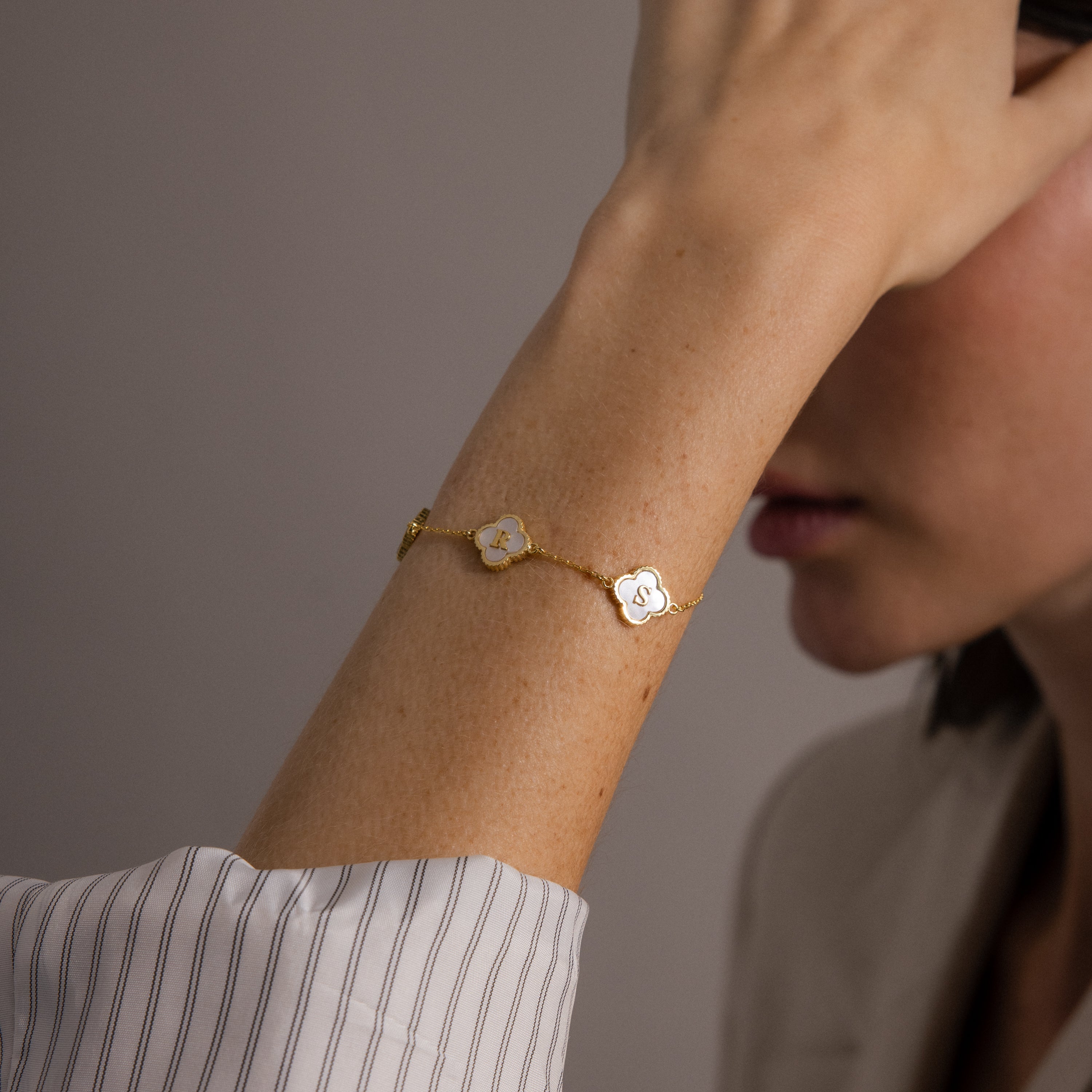 The Pearl Clover Letter Bracelet features two mother of pearl clovers on a woman's wrist with a striped sleeve, hand near face—this personalized bracelet adds chic elegance to any outfit.