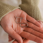 A hand in a green sweater holds two small metal pieces—the Bar & Carabiner Charm Set in White Gold, featuring one U-shaped and one oval-shaped charm.