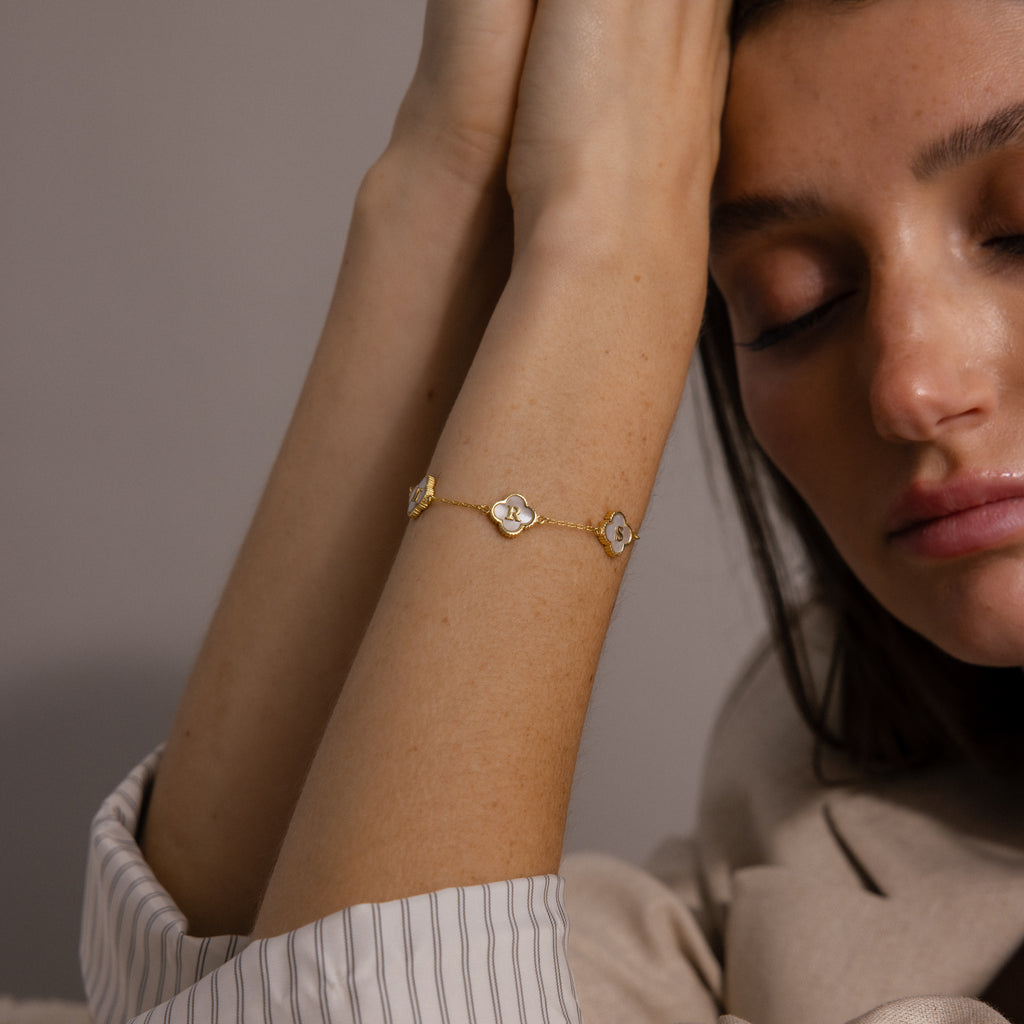 A woman with closed eyes rests her face on her hand, wearing the Pearl Clover Letter Bracelet featuring gold details, mother of pearl clover accents, and delicate floral charms.