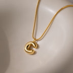The Bubble Letter Necklace in 18K Gold features a chunky, abstract C-shaped pendant and is displayed on a smooth, cream-colored surface.