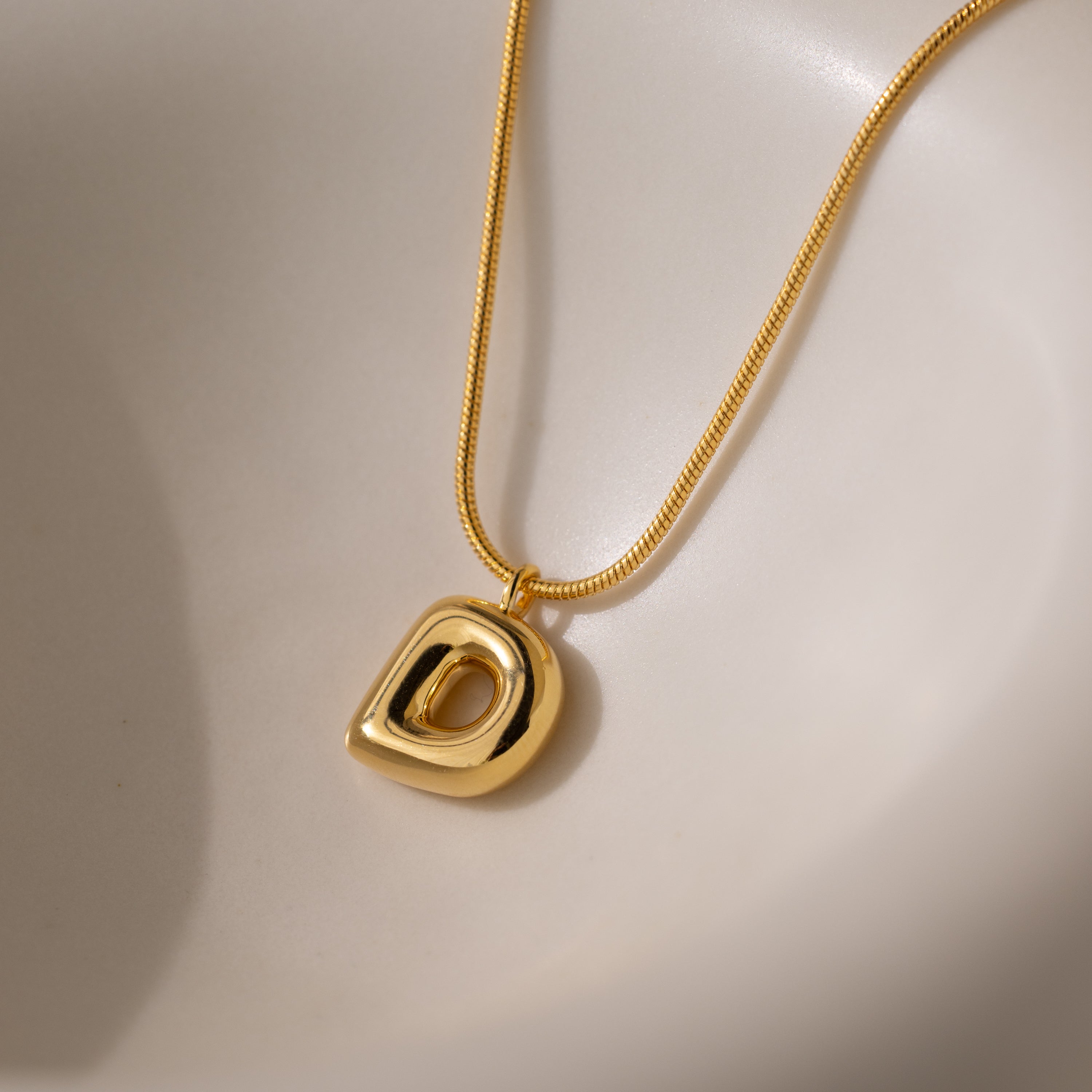 The Bubble Letter Necklace in 18K Gold features a polished D-shaped pendant resting on a smooth, light surface—an eye-catching piece for fans of personalized jewelry.